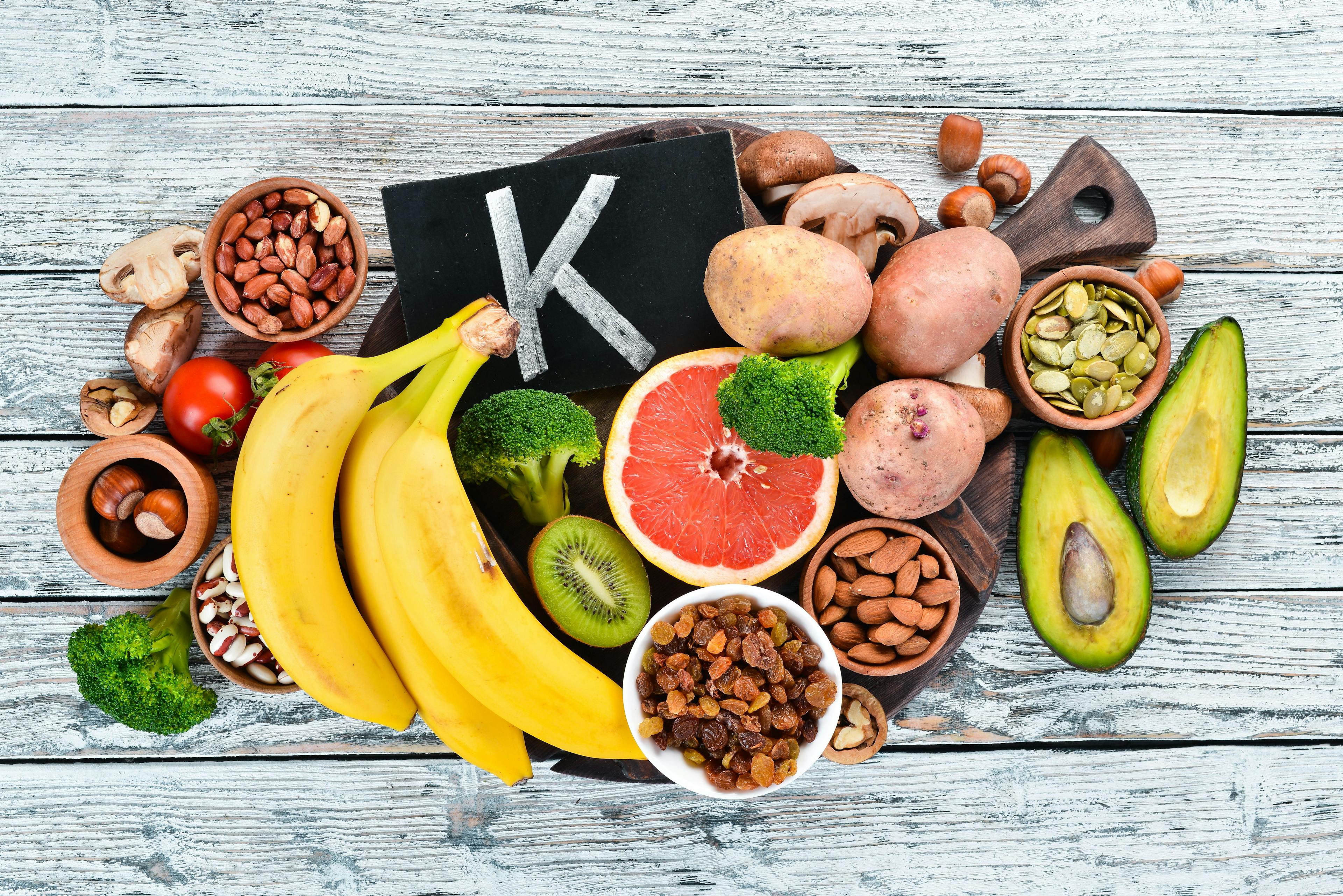 Foods containing natural potassium. K: Potatoes, mushrooms, banana, tomatoes, nuts, beans, broccoli, avocados. Top view. On a white wooden background. | Image Credit: © Yaruniv-Studio
